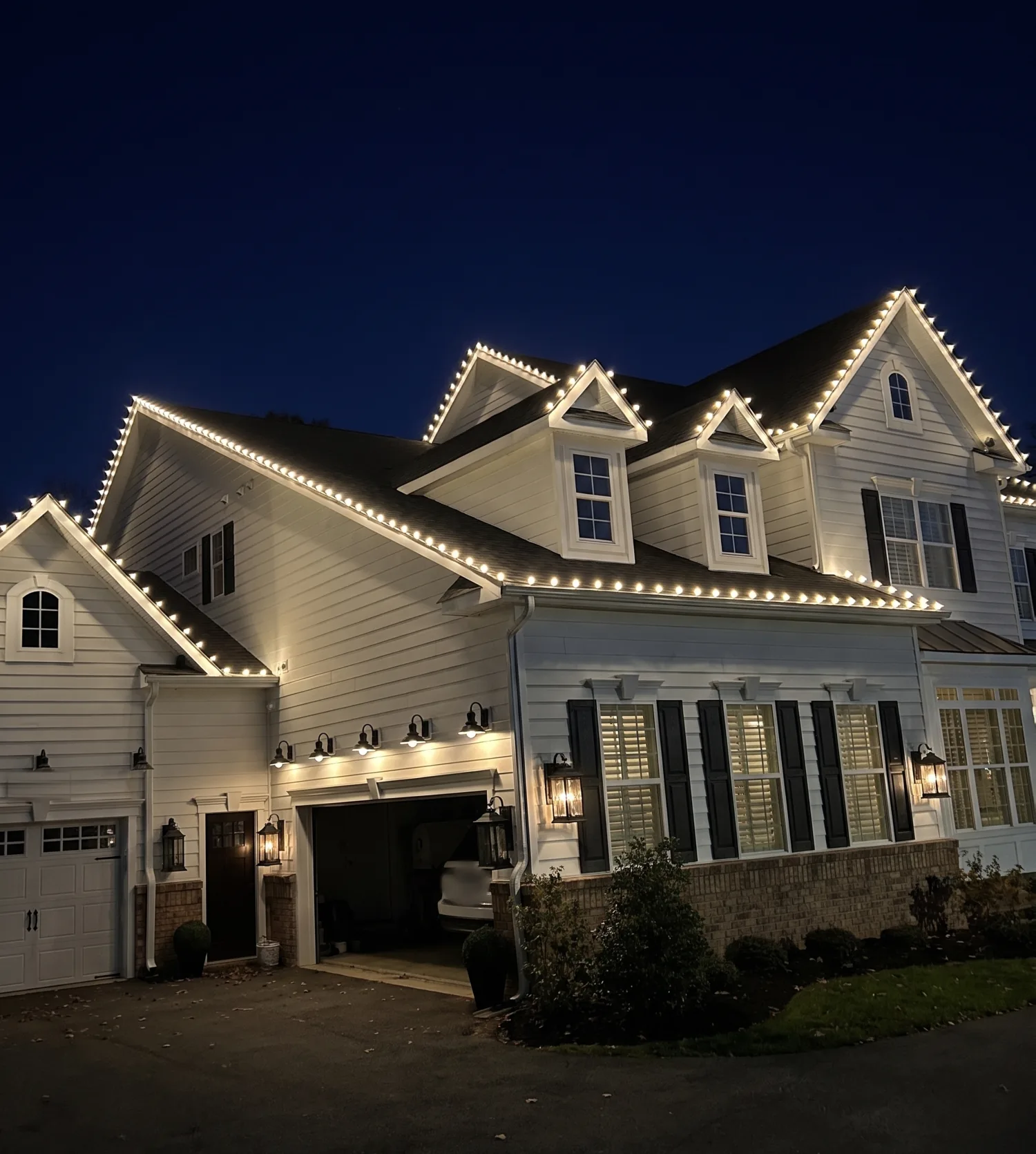 The same white two-story home at night, with warm white lights along its rooflines