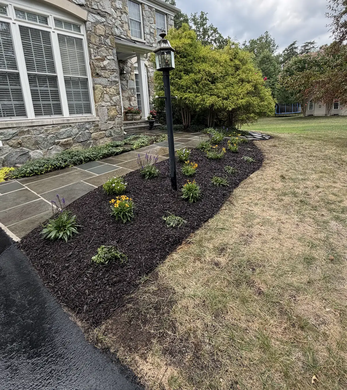 A front bed with dark mulch and new plants round a lamp post, in front of a stone house