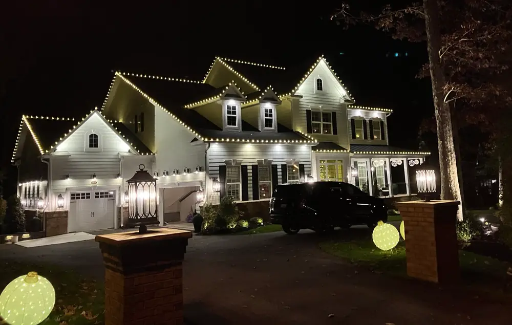 A large white house at night with its roof peaks and garage outlined in warm white lights